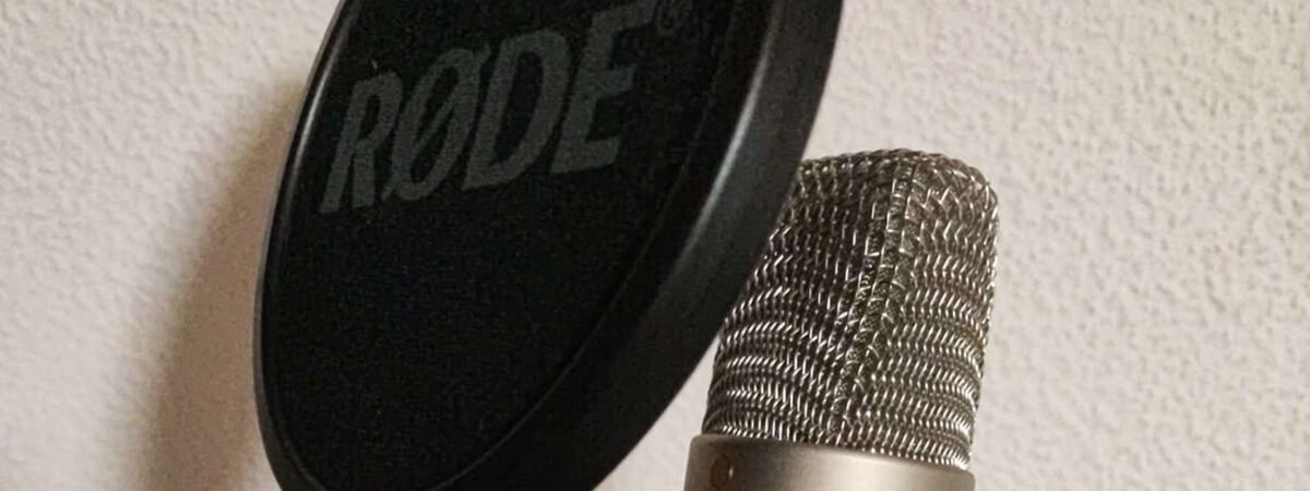 Das silberne Mikrofon der Firma Rode mit einem runden schwarzen Spuckschutz. Rode steht darauf mit weißen Lettern geschrieben. Im Hintergrund ist eine weiße Wand des privaten Tonstudios vom Synchronsprecher Christopher Maximilian Zunke zu sehen.
